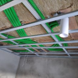 ARKOS - INSTAL ARKADIUSZ STENKA - Zielone rury wentylacyjne w izolacji pod sufitem podwieszanym z widoczną konstrukcją metalową i elementem białej rury.