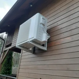 Biała pompa ciepła marki Viessmann zamontowana na brązowej drewnianej elewacji domu, widoczna drabina oparta o ścianę.