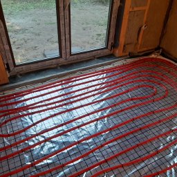 ARKOS - INSTAL ARKADIUSZ STENKA - Czerwone rury ogrzewania podłogowego ułożone na srebrnej folii refleksyjnej z siatką, widoczne surowe ściany z izolacją, okno z widokiem na zewnątrz.