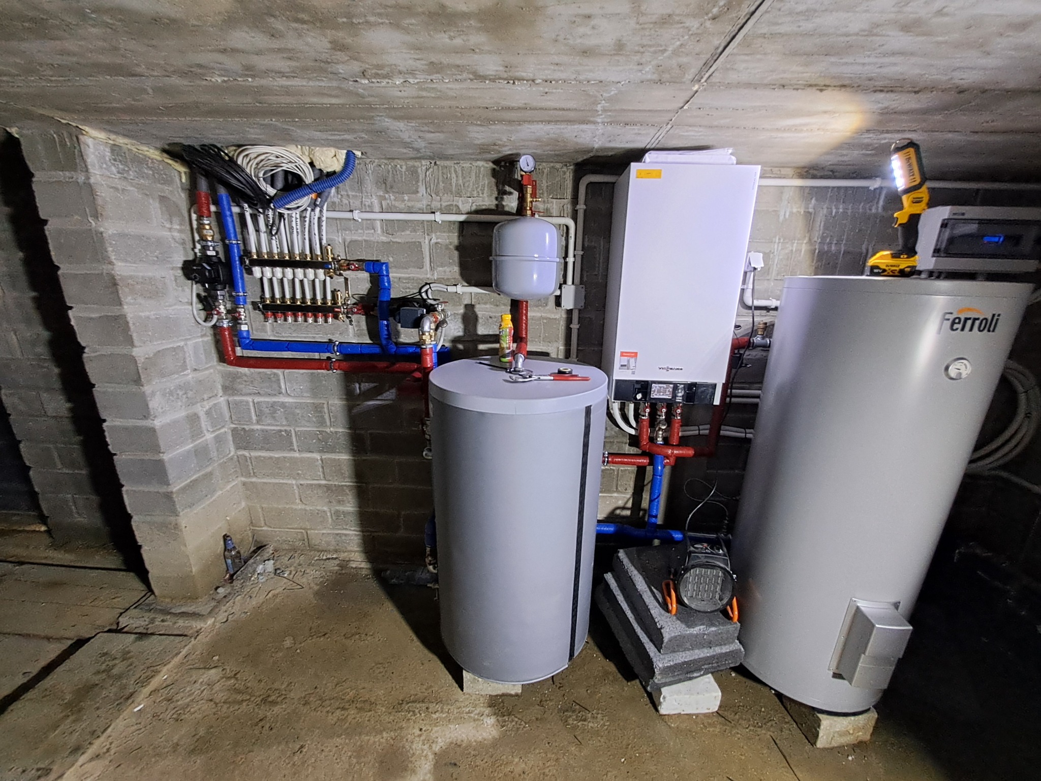 Kotłownia w piwnicy z piecem Viessmann, zasobnikiem Ferolli, rozdzielaczem, rurami w kolorze czerwonym i niebieskim, oraz lampą roboczą DeWalt na zasobniku.
