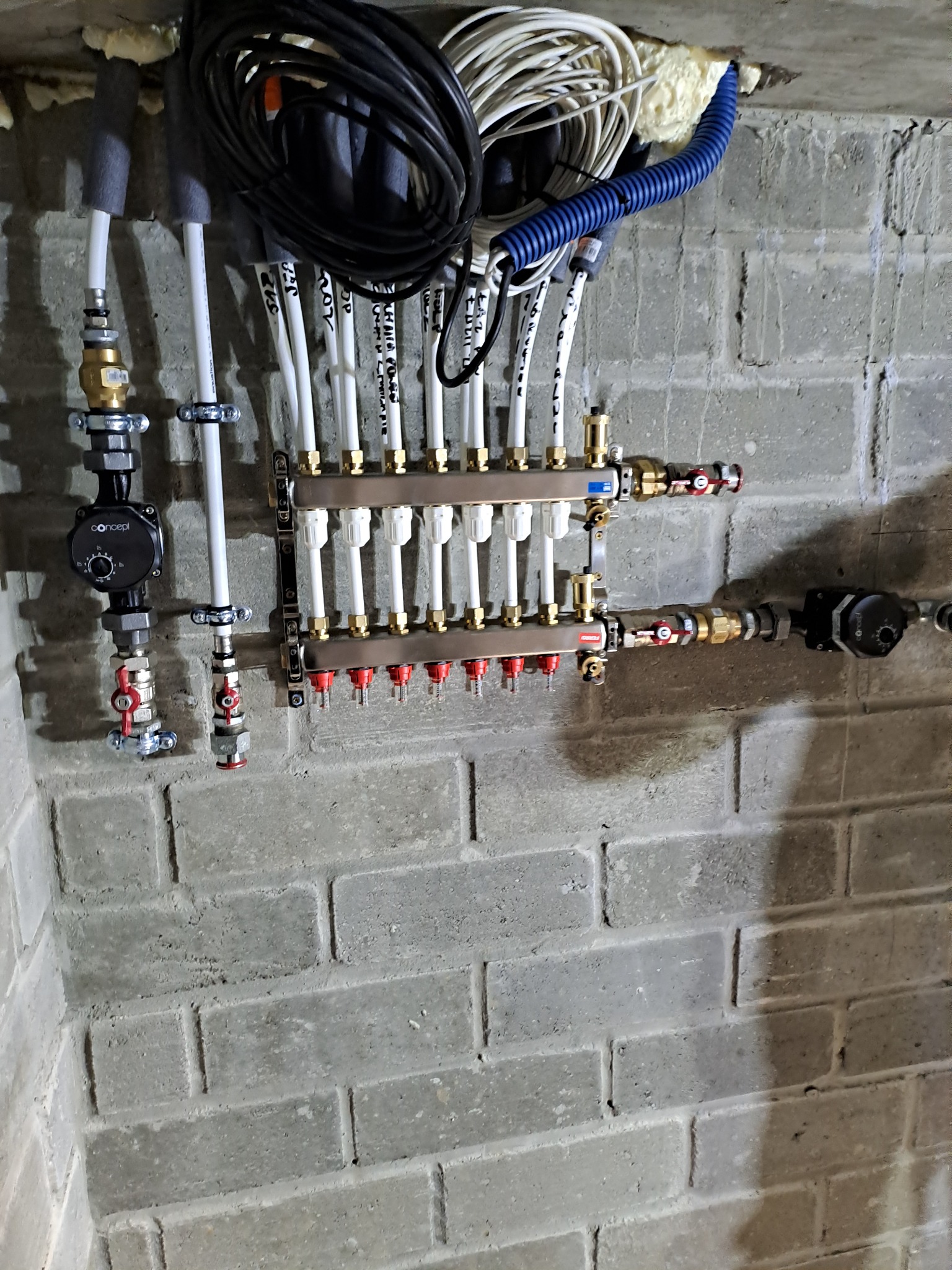 Rozdzielacz ogrzewania podłogowego z rurami PEX i pompą obiegową zamontowany na szarej, surowej ścianie z cegieł. Widoczne przewody elektryczne i izolacja piankowa.