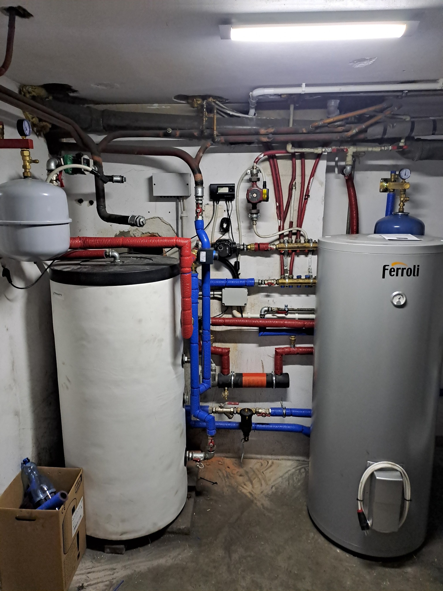 Dwa zbiorniki na wodę, biały i szary Ferroli, połączone rurami w piwnicy z betonową podłogą i sufitem pod jarzeniówką.