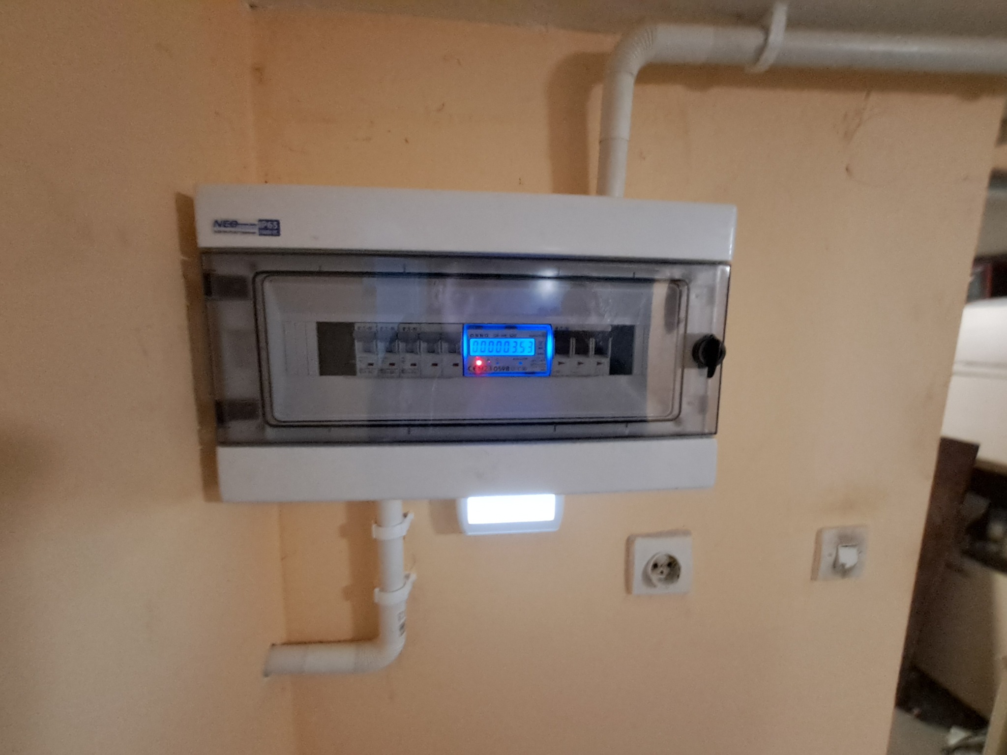 Szczegółowe ujęcie skrzynki rozdzielczej z licznikiem energii elektrycznej, zamontowanej na beżowej ścianie, z widocznymi przewodami i gniazdkami elektrycznymi.