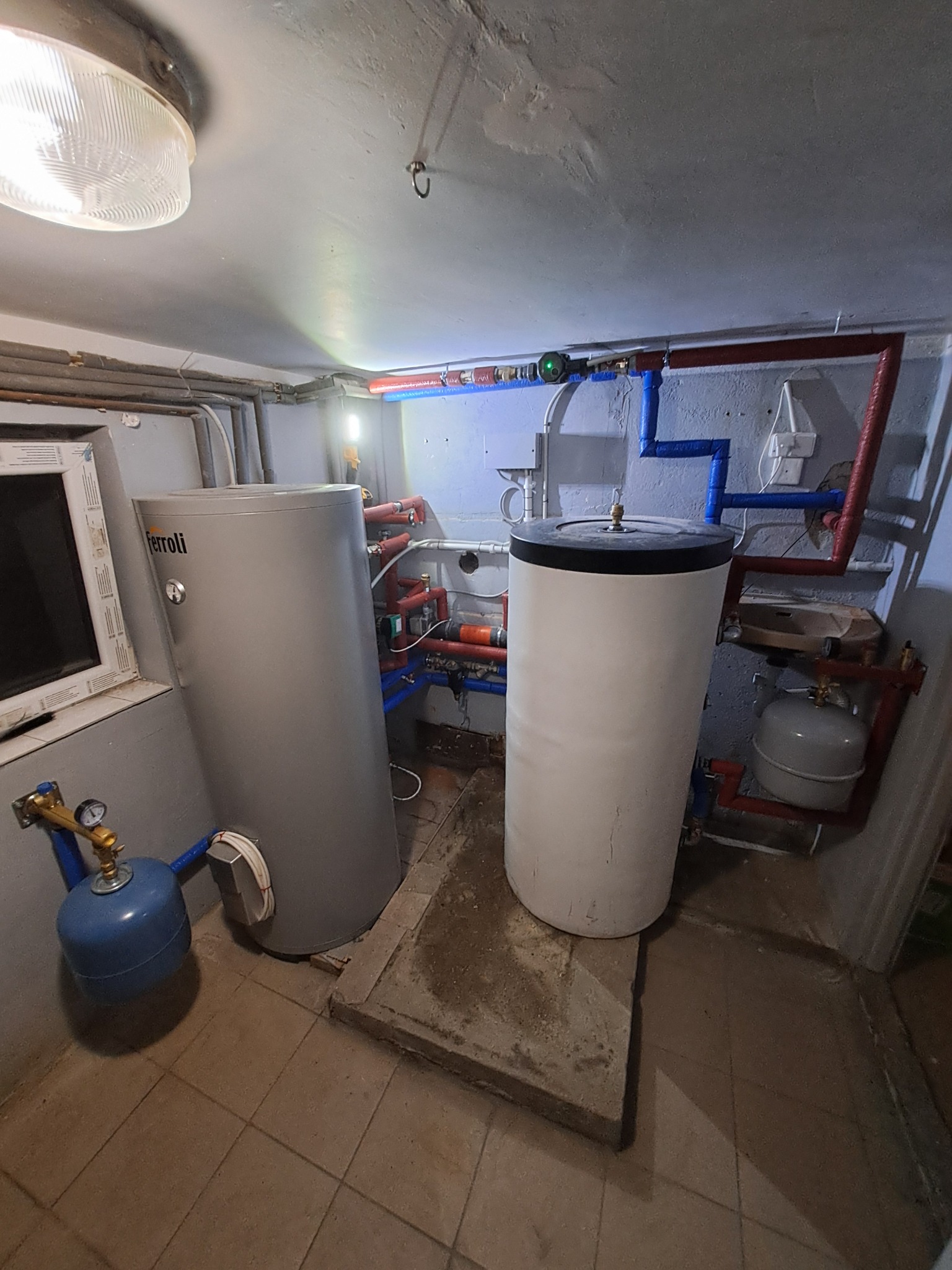 Dwa zbiorniki na wodę, jeden szary marki Ferroli, drugi biały, połączone rurami w pomieszczeniu kotłowni z widocznymi elementami instalacji grzewczej.