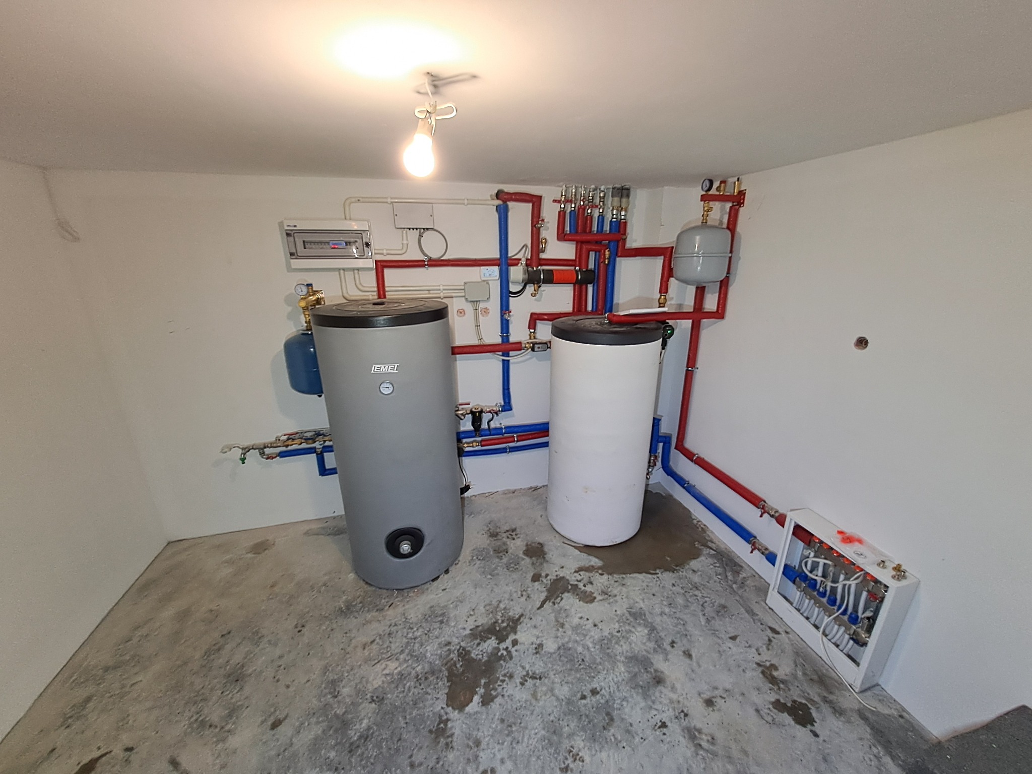 Dwa zbiorniki akumulacyjne ciepła (szary i biały) w kotłowni z instalacją hydrauliczną w kolorach czerwonym i niebieskim, widoczne również rozdzielacze i osprzęt kotłowy, betonowa posadzka.