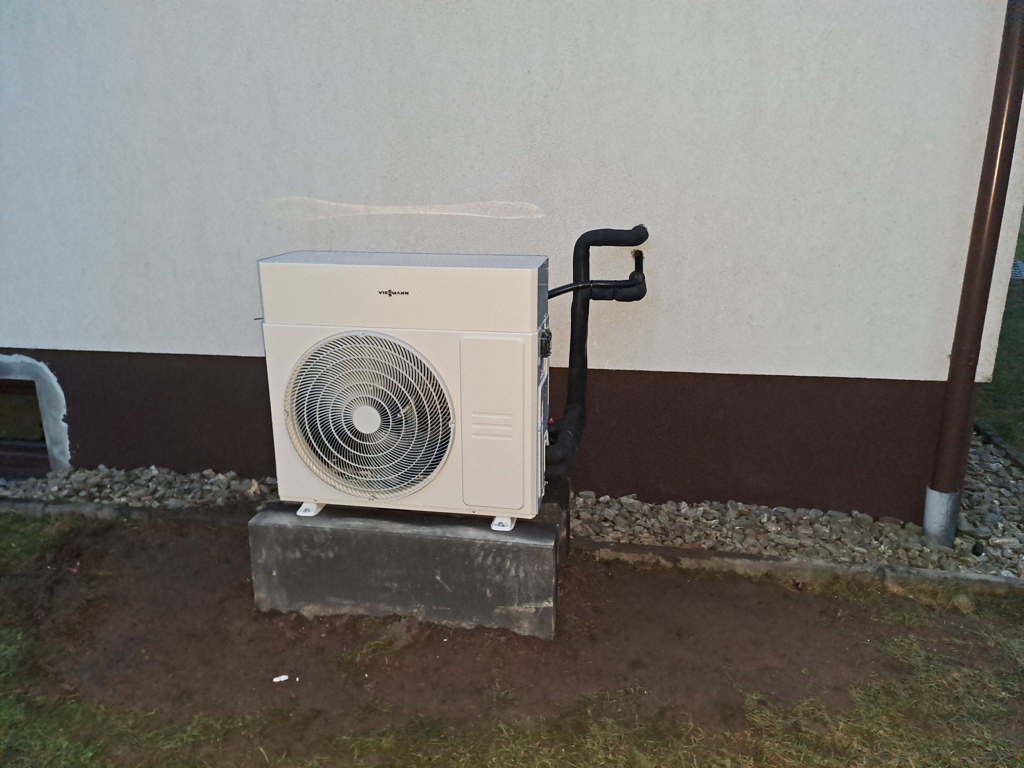 Zamontowana zewnętrzna jednostka pompy ciepła Viessmann na betonowym fundamencie przy ścianie budynku z widocznymi rurami izolowanymi termicznie.