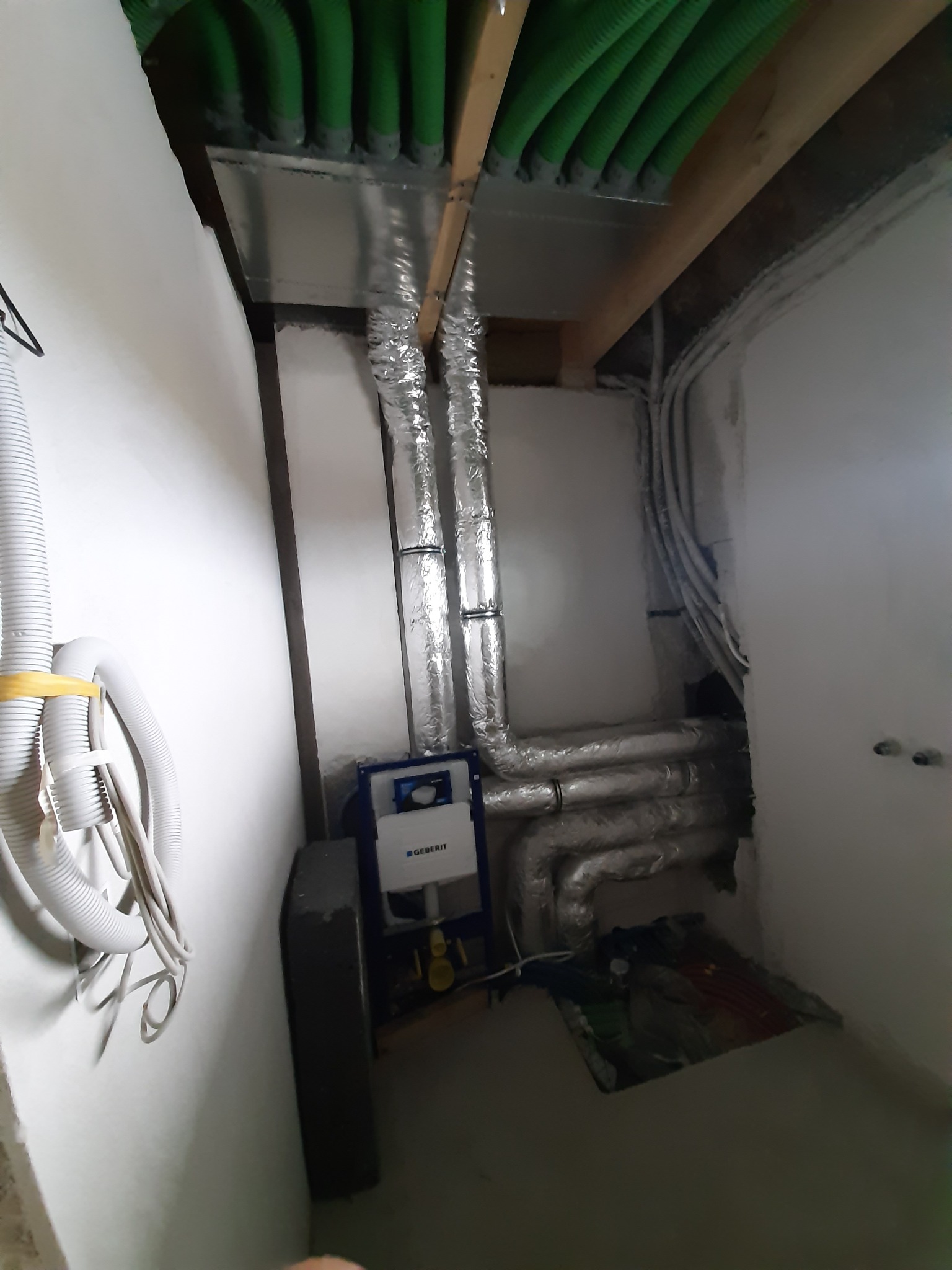 Instalacja podtynkowa Geberit widoczna w surowym stanie, rury wentylacyjne w izolacji aluminiowej, zielone rury instalacyjne pod sufitem, przewody elektryczne w ścianie.