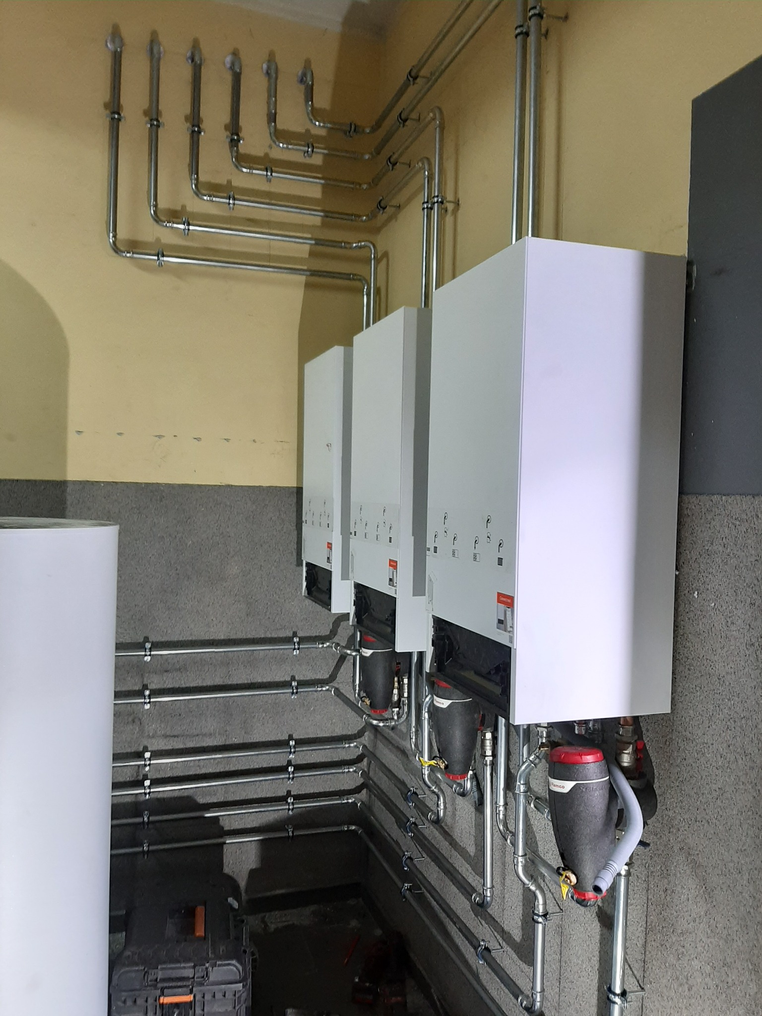 Trzy identyczne białe piece centralnego ogrzewania zamontowane na szarej ścianie, widoczne liczne rury instalacji grzewczej i filtrów, skrzynka narzędziowa na podłodze.