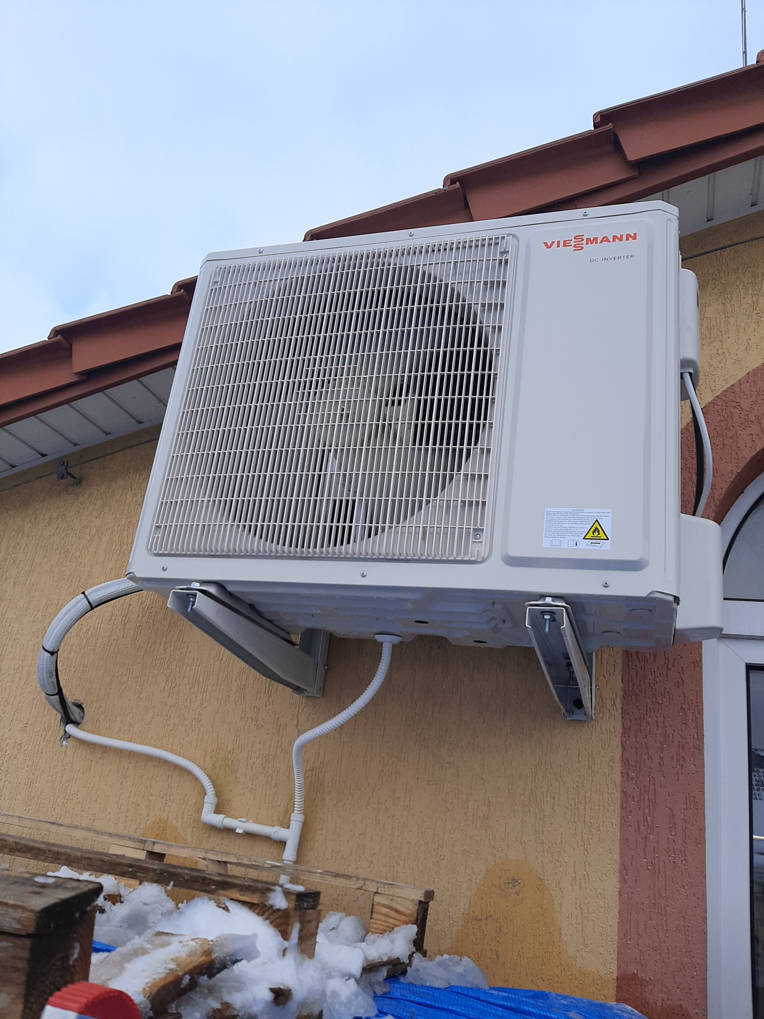 Zewnętrzna jednostka klimatyzacji Viessmann DC Inverter zamontowana na elewacji budynku, widoczne rury instalacyjne i fragment dachu z dachówką.