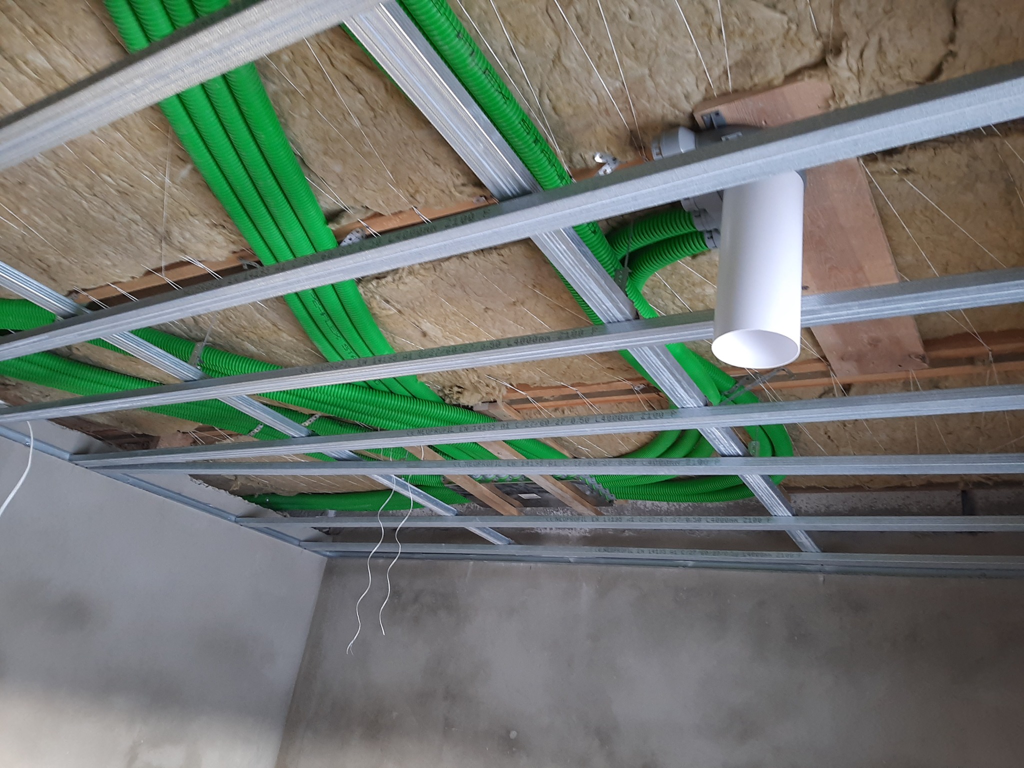 Zielone rury wentylacyjne w izolacji pod sufitem podwieszanym z widoczną konstrukcją metalową i elementem białej rury.