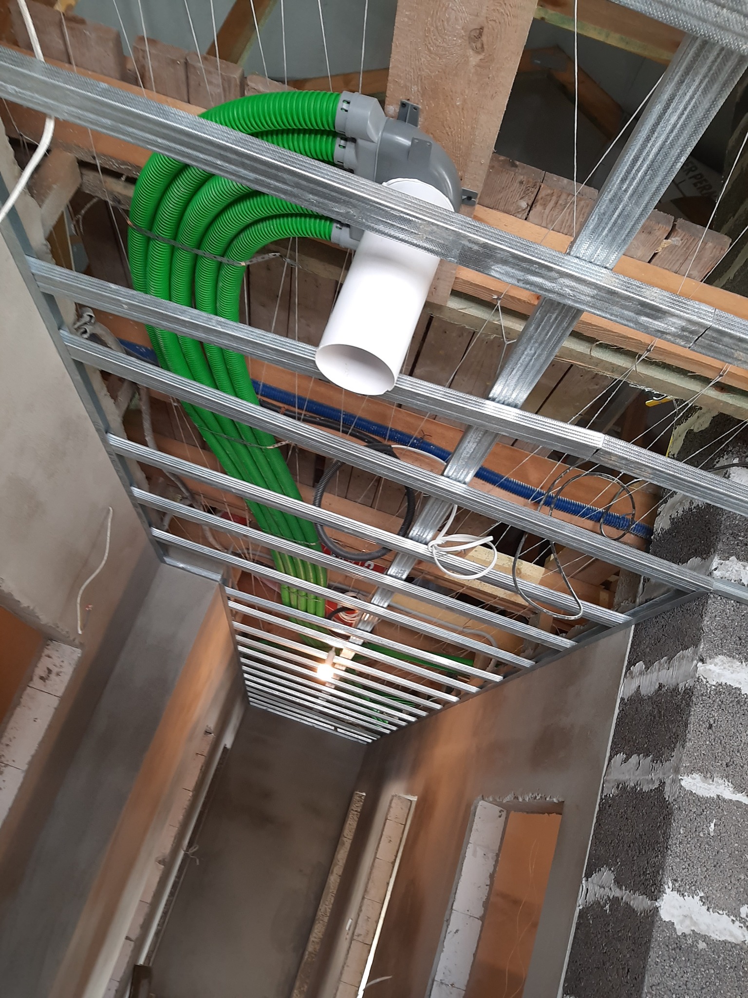 Montaż instalacji wentylacyjnej z użyciem zielonych, elastycznych rur w budynku w stanie surowym w Luzinie, widok z dołu na konstrukcję podwieszanego sufitu.