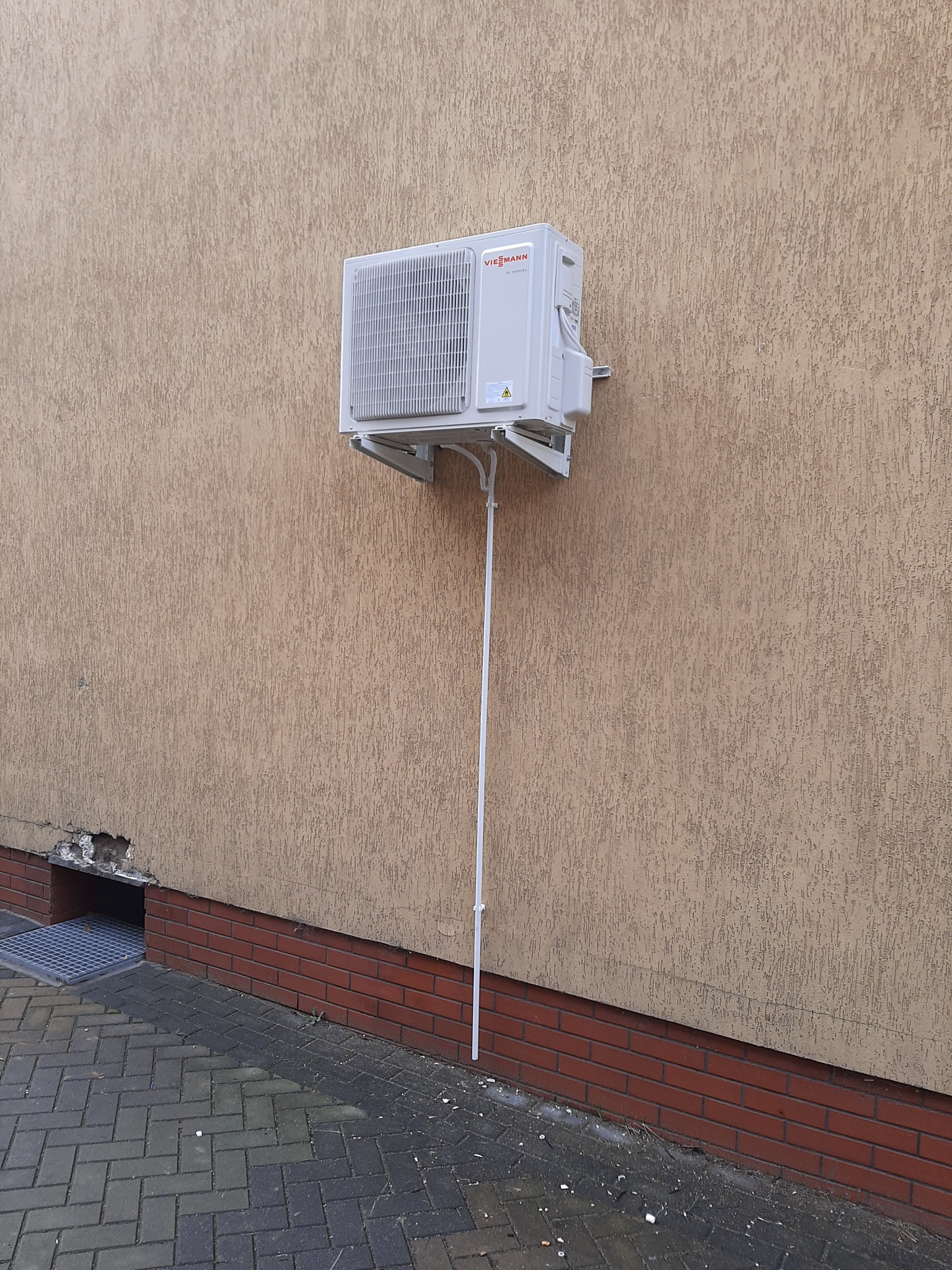 Biała jednostka zewnętrzna pompy ciepła Viessmann zamontowana na beżowej elewacji budynku z widoczną instalacją odprowadzającą skropliny.