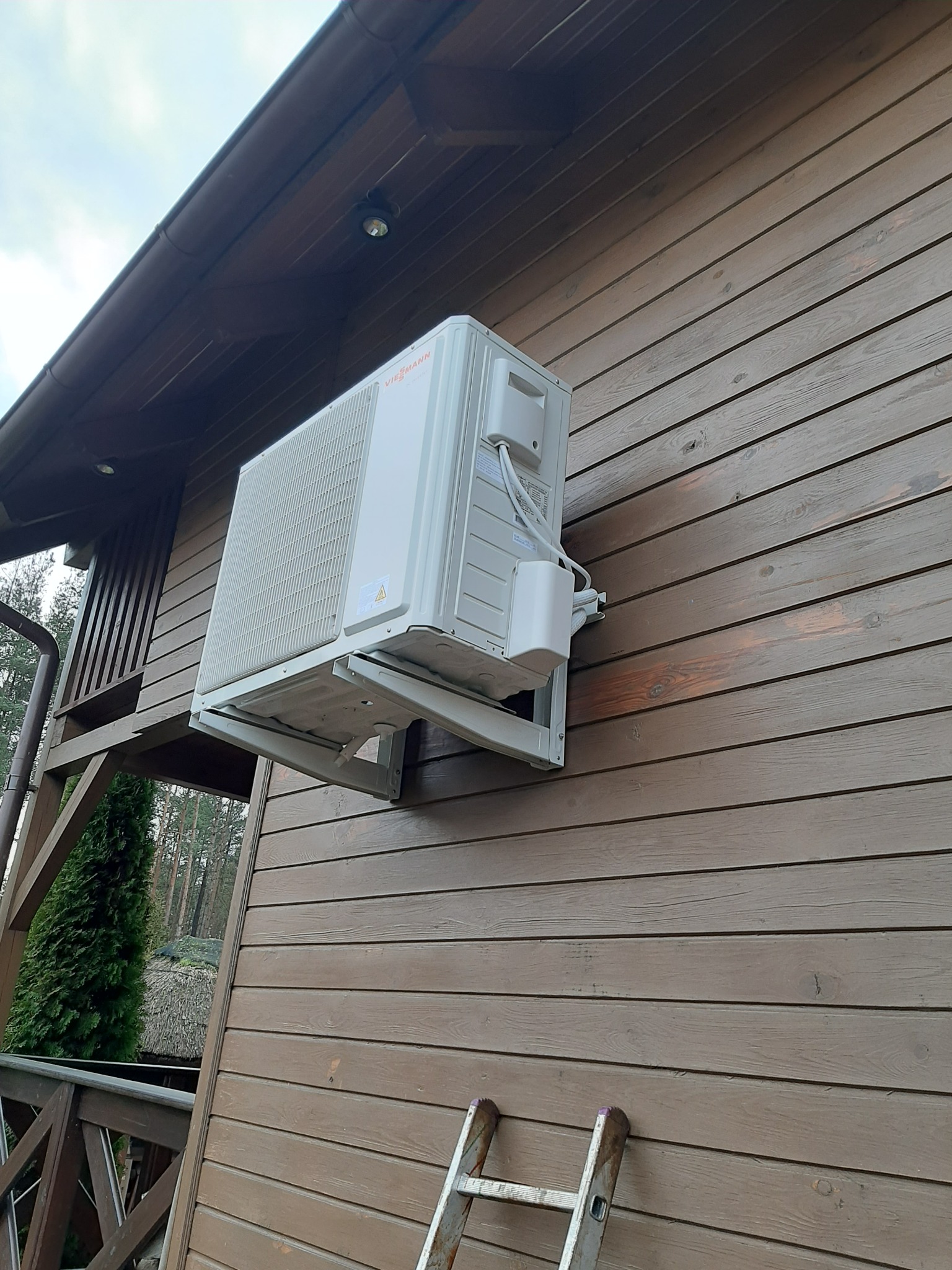 Biała pompa ciepła marki Viessmann zamontowana na brązowej drewnianej elewacji domu, widoczna drabina oparta o ścianę.