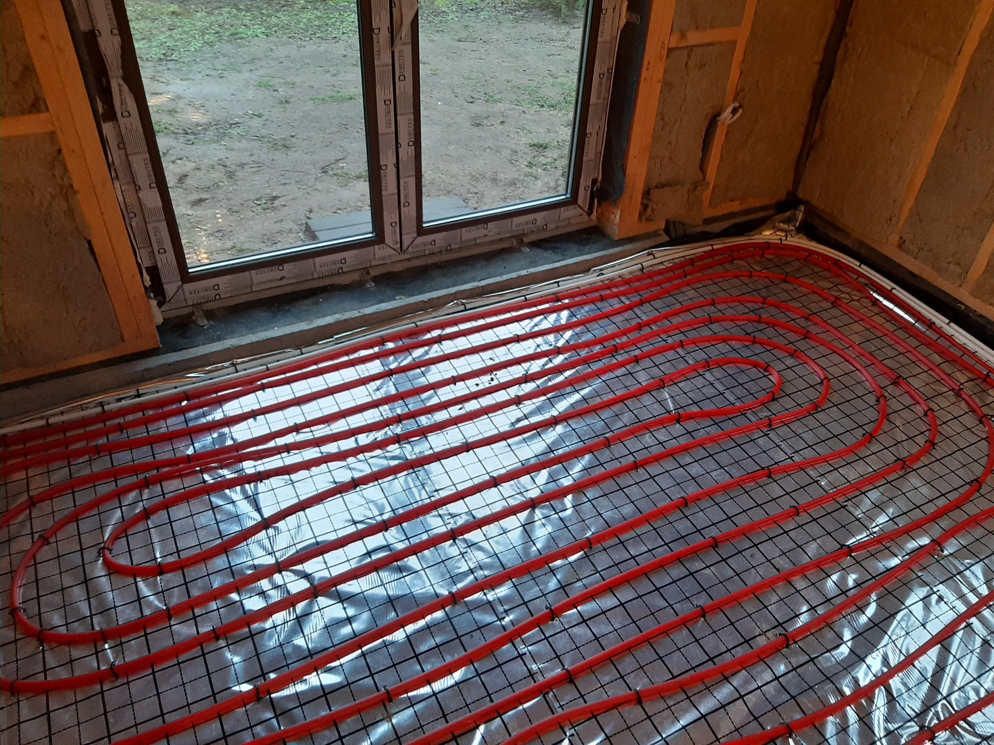 Czerwone rury ogrzewania podłogowego ułożone na srebrnej folii refleksyjnej z siatką, widoczne surowe ściany z izolacją, okno z widokiem na zewnątrz.