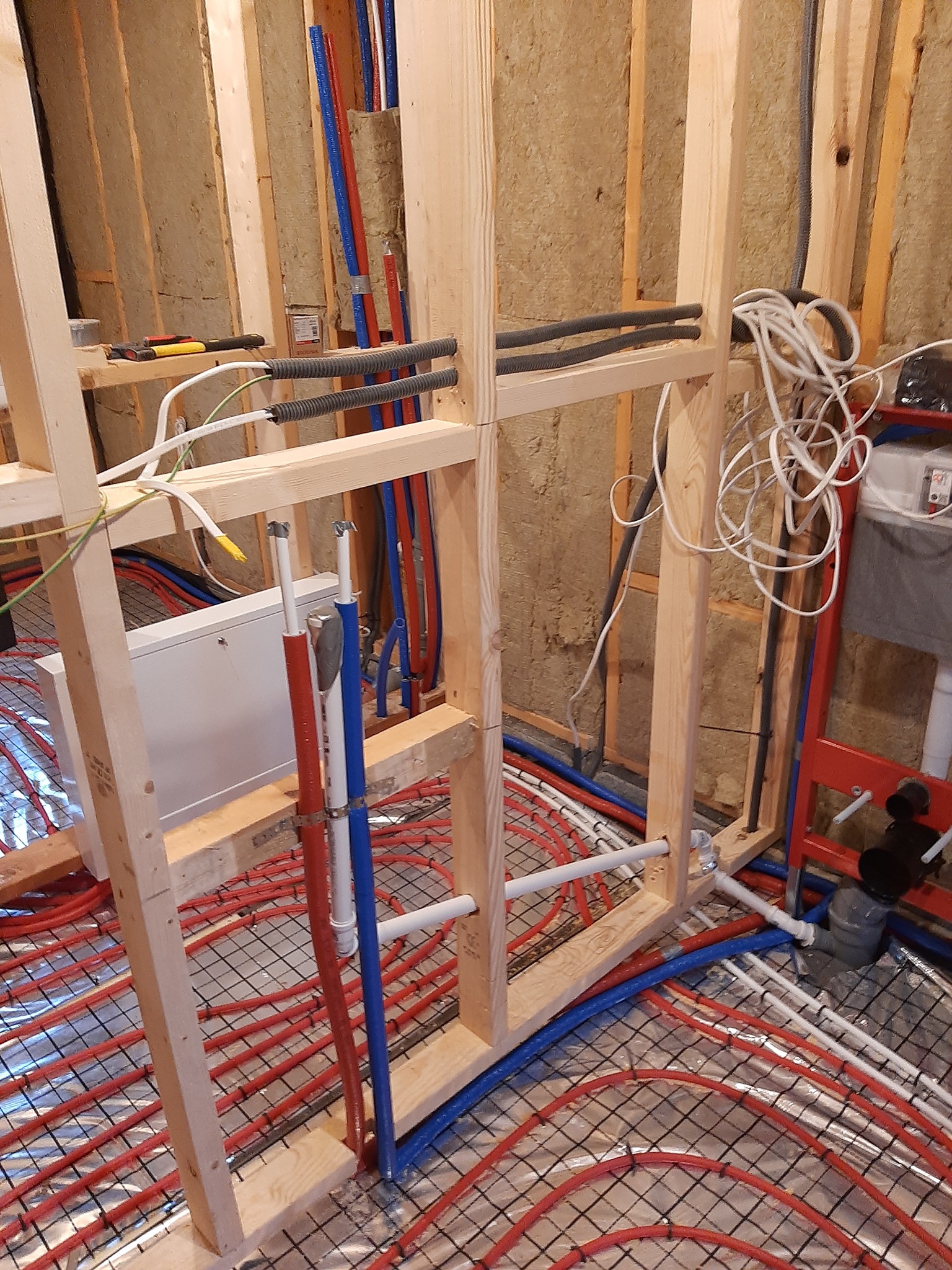 Montaż instalacji ogrzewania podłogowego: widoczne czerwone rury na metalowej siatce, drewniana konstrukcja ściany z izolacją, oraz pionowe rury hydrauliczne w kolorze niebieskim i czerwonym.