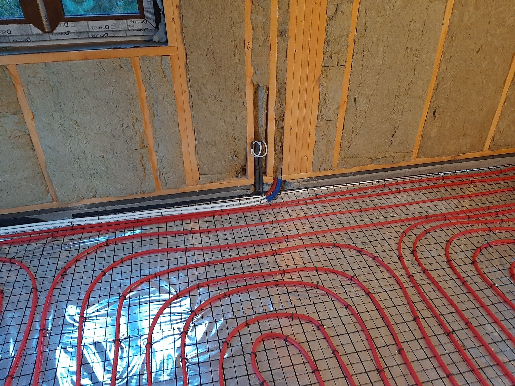 Montaż czerwonych rur ogrzewania podłogowego na metalowej siatce, widoczne ocieplone ściany z wełny mineralnej i pionowe rury instalacyjne w ścianie.