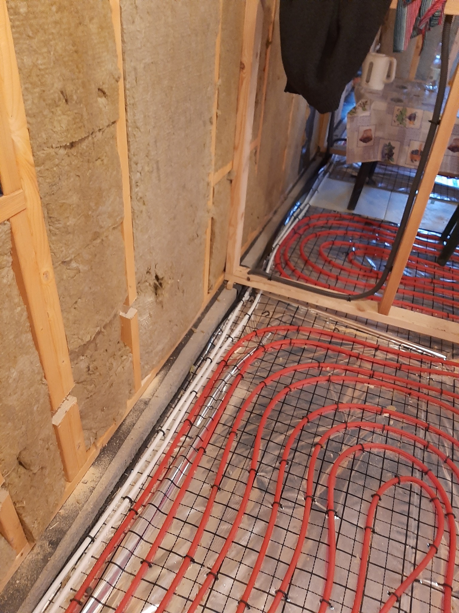 Instalacja ogrzewania podłogowego w trakcie budowy, widoczne czerwone rury na folii aluminiowej i izolacja ścian wełną mineralną.