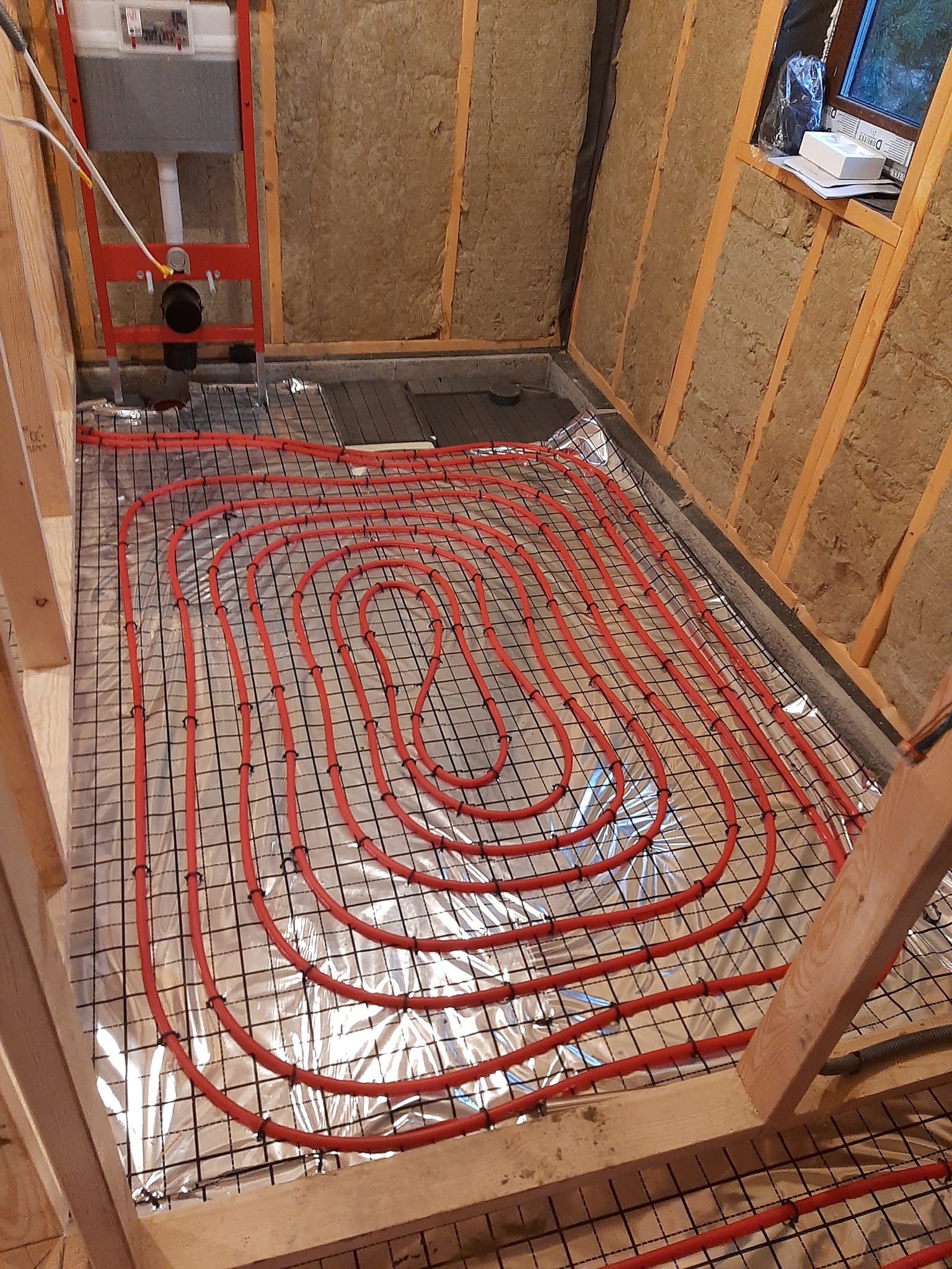 Instalacja czerwonych rur ogrzewania podłogowego na folii aluminiowej, zamocowanych do metalowej siatki w pomieszczeniu z ocieplonymi ścianami i stelażem podtynkowym WC.