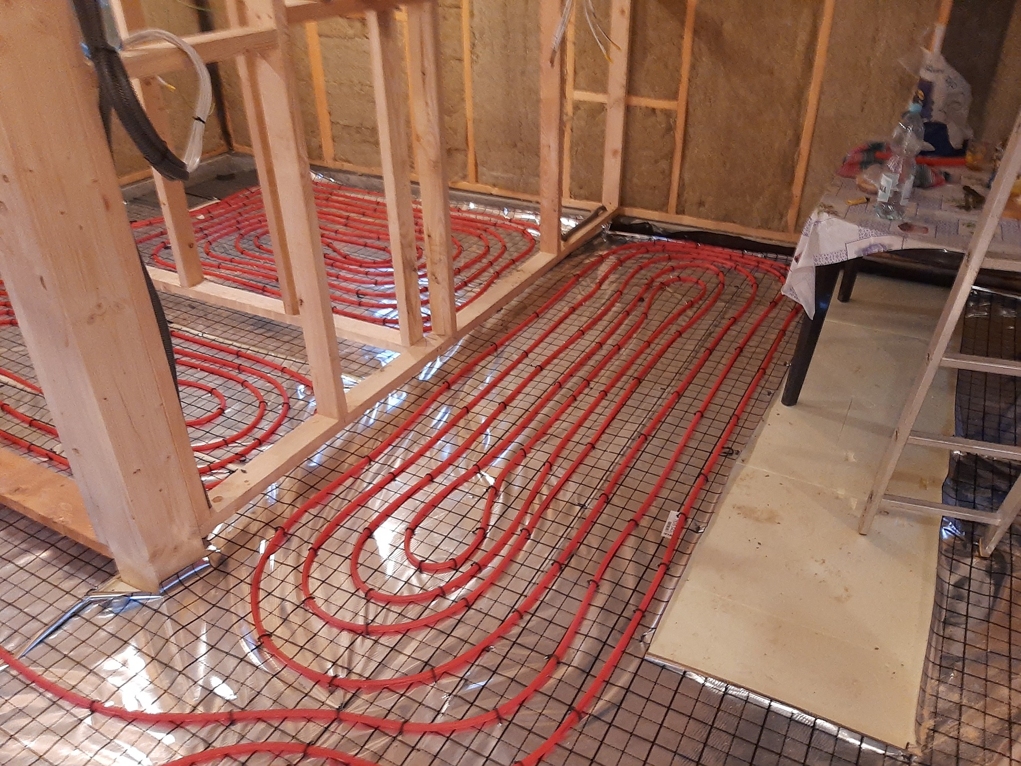 Montaż czerwonych rur ogrzewania podłogowego na metalowej siatce, widoczne drewniane elementy konstrukcji ścian i fragment stołu z narzędziami.