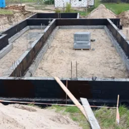 Zalane fundamenty budynku z wystającymi prętami zbrojeniowymi, zabezpieczone czarną izolacją. Część bloczków czeka na ułożenie w centralnej części wykopu.