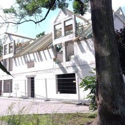 Viking_Dom Adrian Wiczling - Surowy stan domu z białego betonu komórkowego, z widoczną konstrukcją dachu i niezamontowanymi oknami, ogrodzony tymczasowym płotem, na tle zieleni.