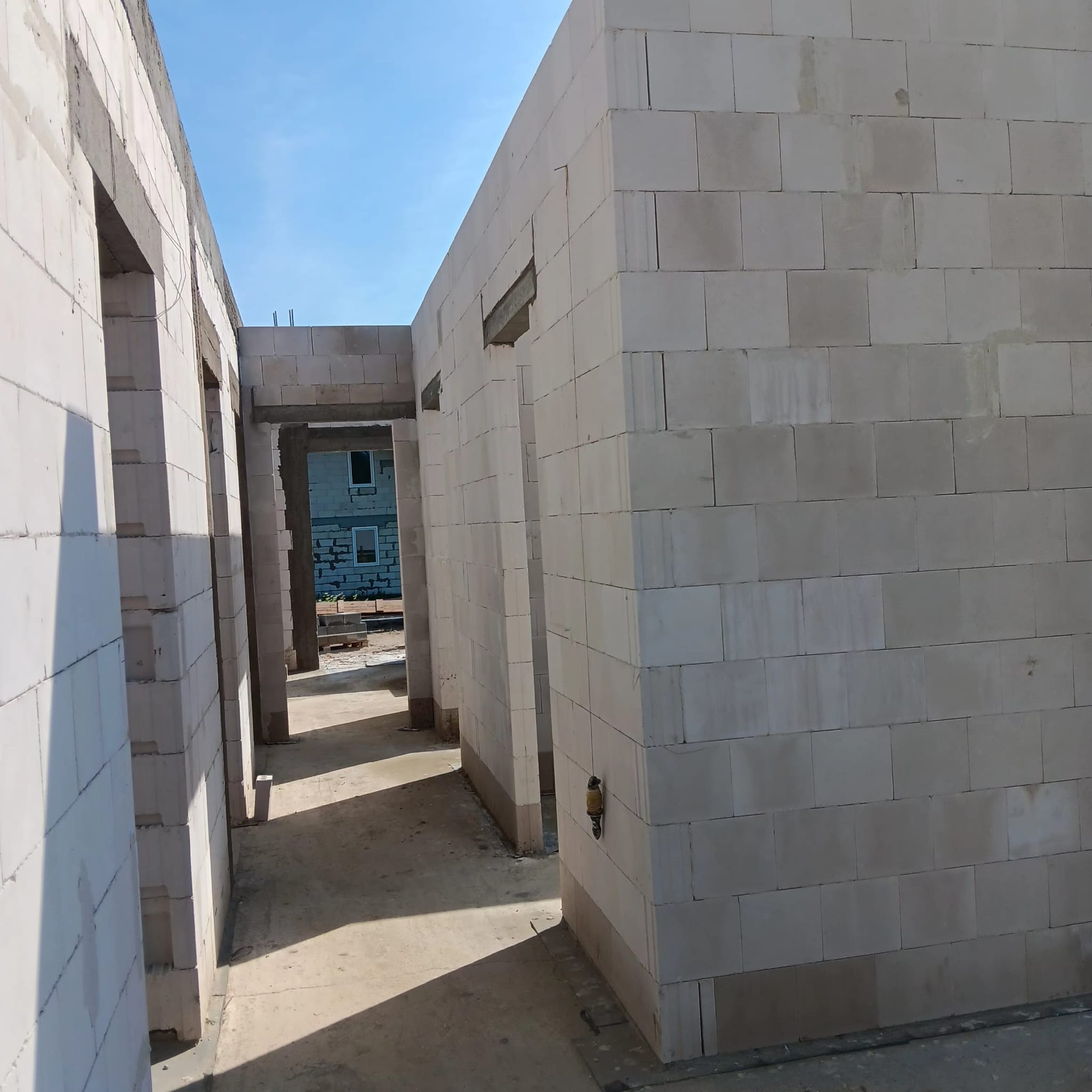 Wnętrze domu w trakcie budowy, widoczne ściany z białych bloczków, otwory na drzwi, surowy stan. Na zewnątrz widoczny budynek w tle, błękitne niebo.