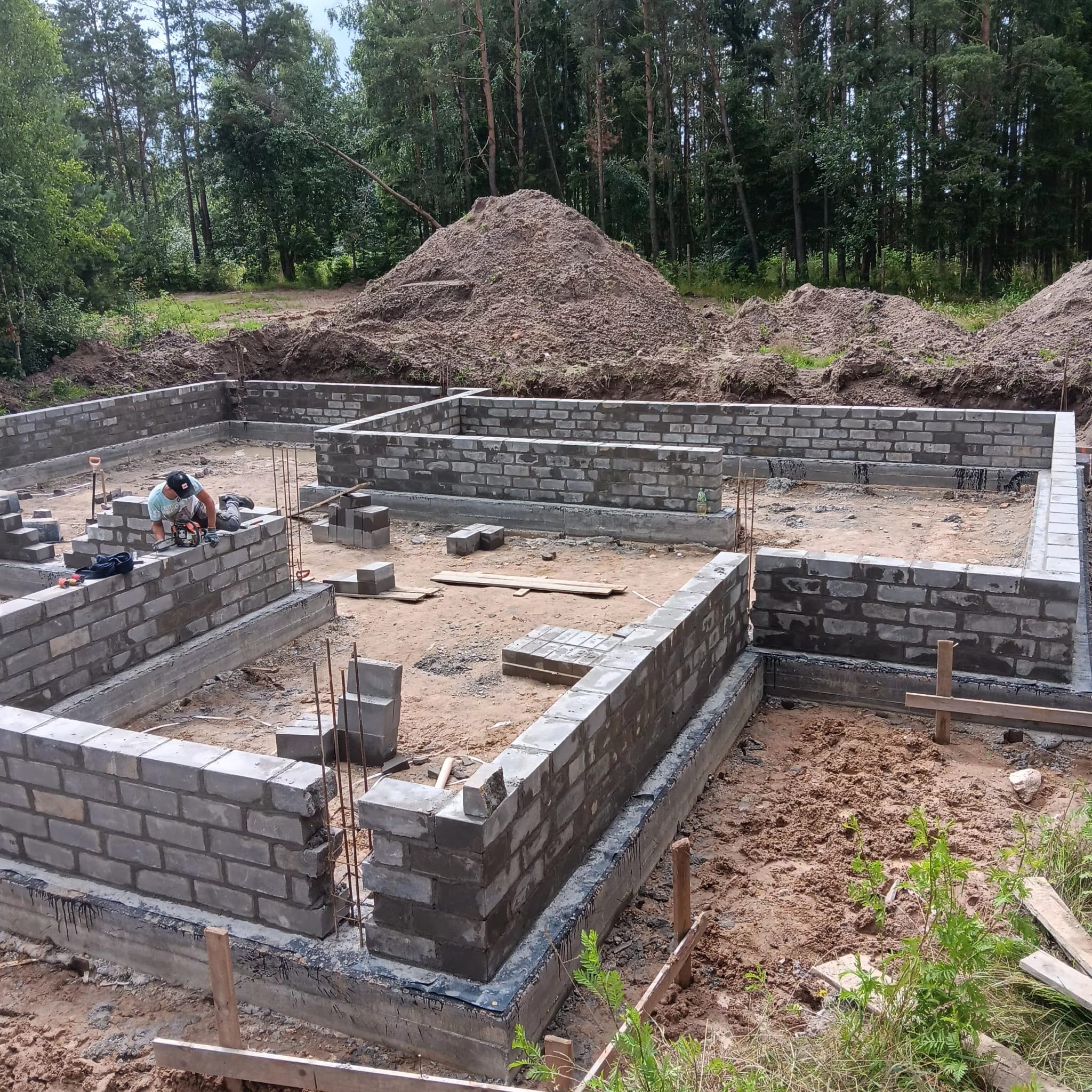 Budowa fundamentów z bloczków betonowych na działce z drzewami w tle. Murarz przy pracy, widoczne narzędzia i materiały budowlane. Etap wznoszenia ścian fundamentowych.