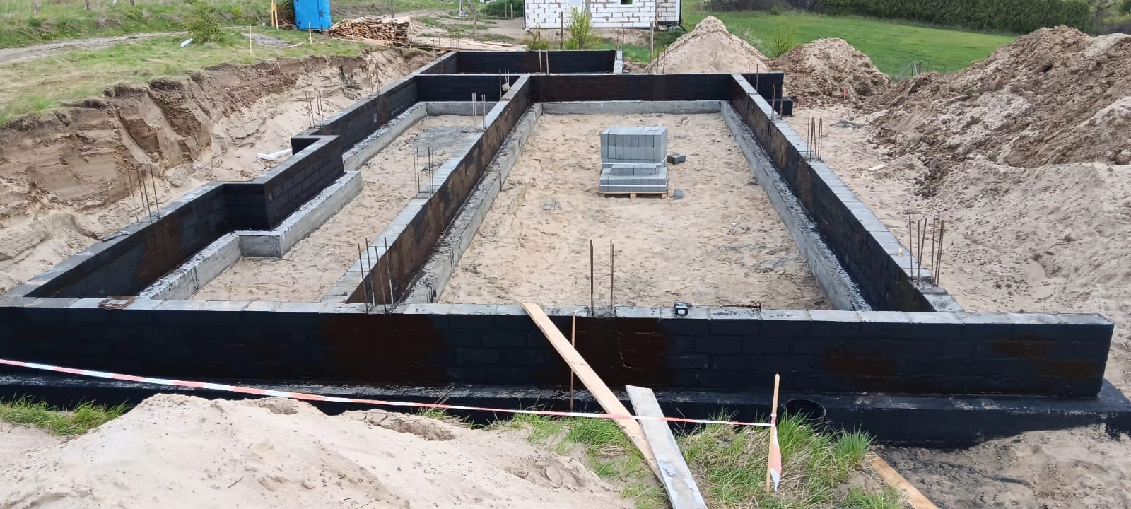 Zalane fundamenty budynku z wystającymi prętami zbrojeniowymi, zabezpieczone czarną izolacją. Część bloczków czeka na ułożenie w centralnej części wykopu.