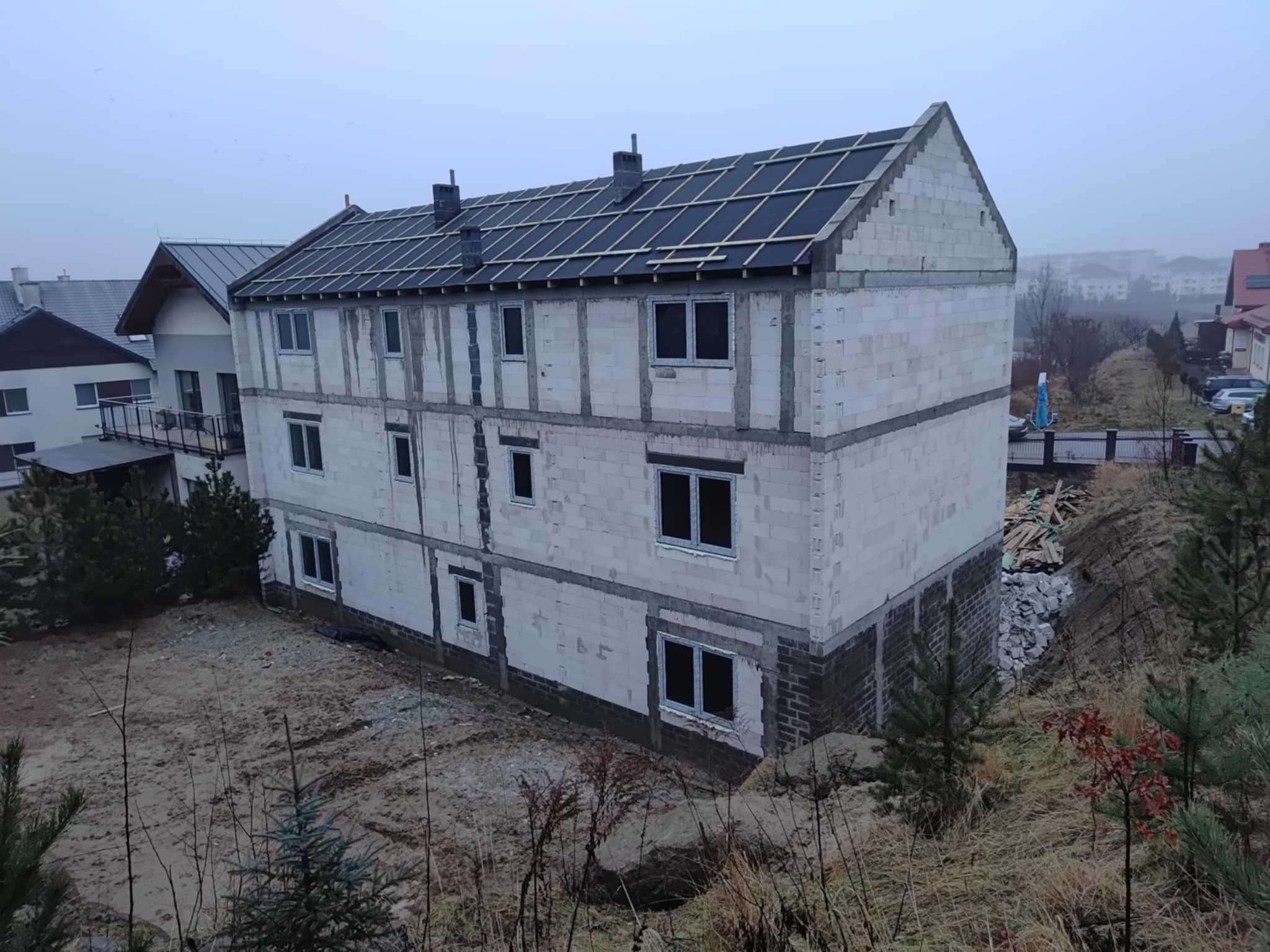 Trzykondygnacyjny dom w stanie surowym otwartym z oknami, z czarnym dachem w trakcie budowy, na tle pochmurnego nieba i zabudowy jednorodzinnej.