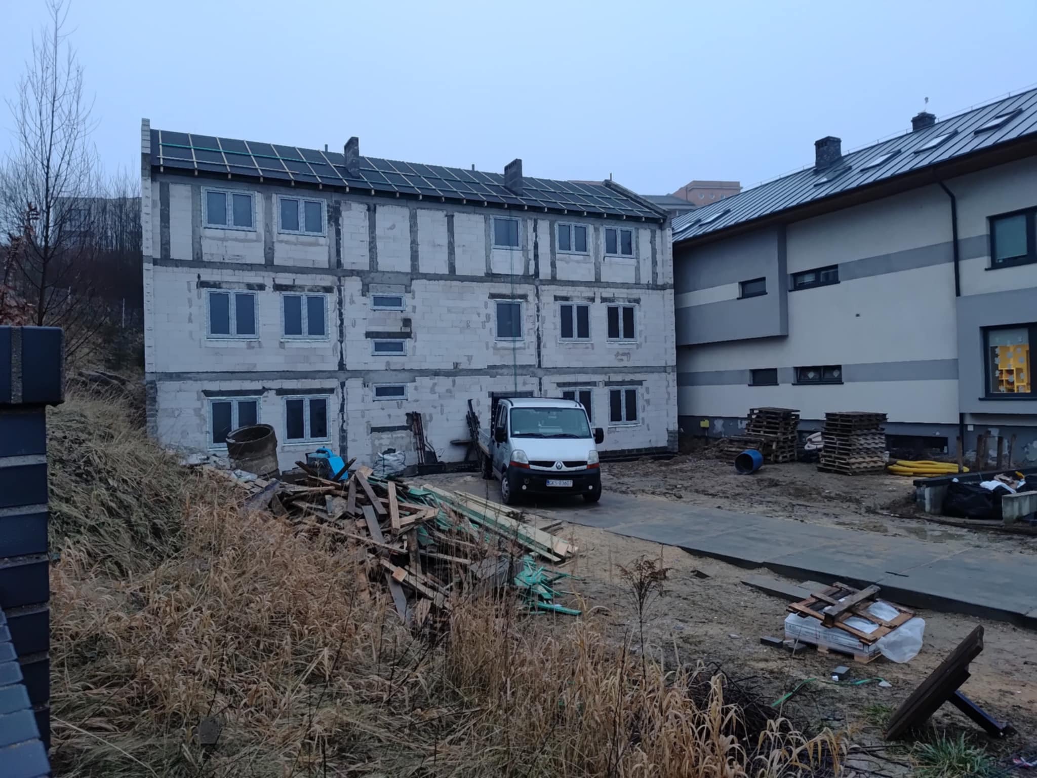 Trzy kondygnacyjny budynek w stanie surowym otwartym z częściowo wykonanym dachem, na pierwszym planie sterta drewna, samochód dostawczy i otoczenie placu budowy.