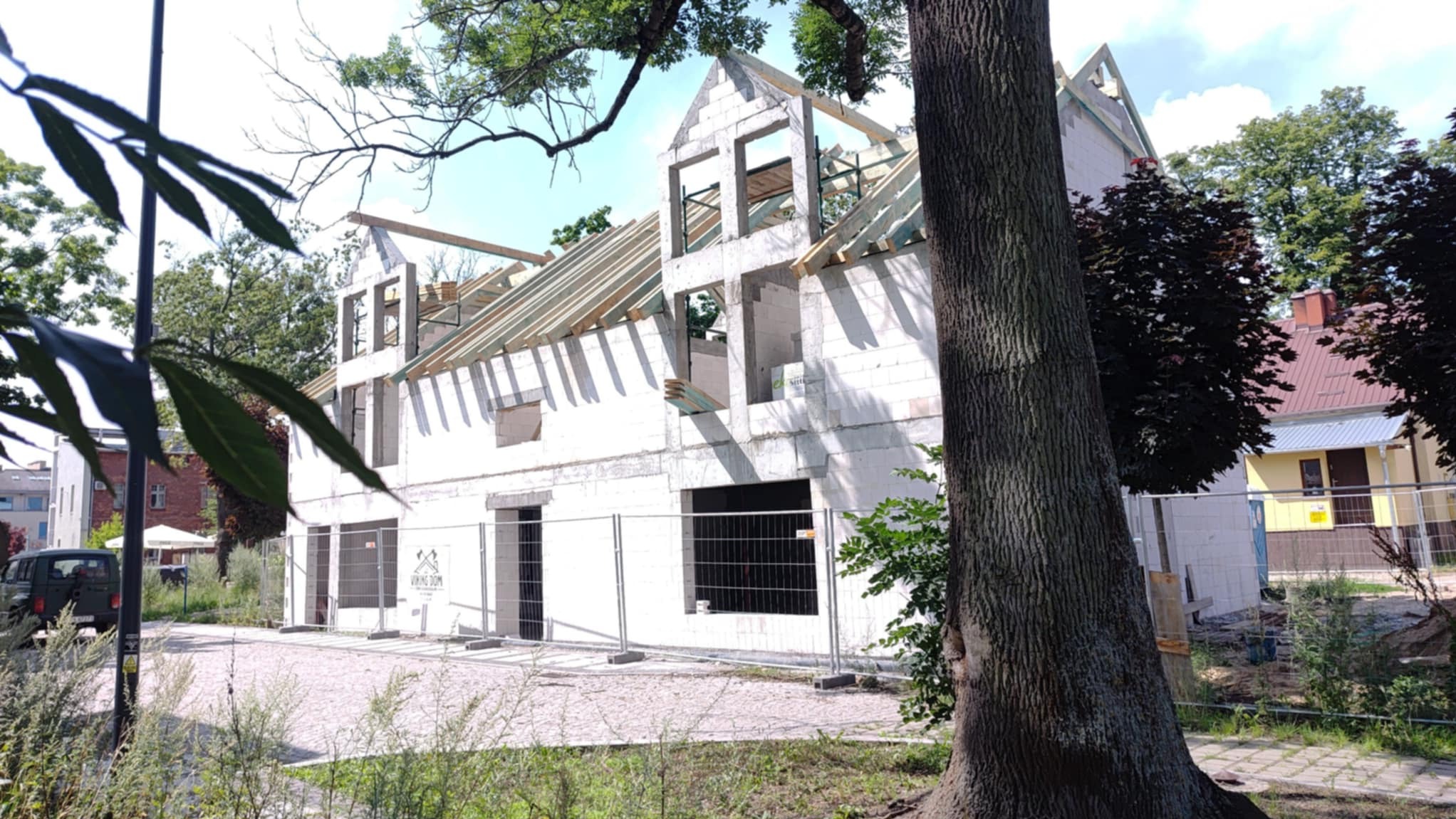 Surowy stan domu z białego betonu komórkowego, z widoczną konstrukcją dachu i niezamontowanymi oknami, ogrodzony tymczasowym płotem, na tle zieleni.