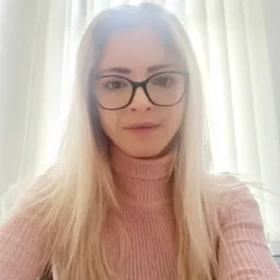 Portret kobiety w okularach i różowym swetrze na tle okna z białymi zasłonami.