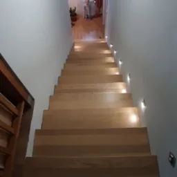 Widok z g&oacute;ry na drewniane schody z subtelnym oświetleniem punktowym wzdłuż ściany, w tle widoczna bawiąca się dziewczynka w pokoju.