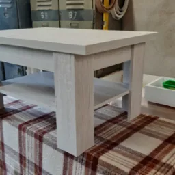 Stolik kawowy z jasnego drewna o widocznej strukturze, z dodatkową półką, ustawiony na pledzie w kratę w warsztacie.