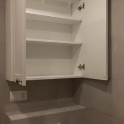 Biała szafka łazienkowa wisząca z otwartymi drzwiczkami, widoczne trzy półki, złoty uchwyt, szare płytki na ścianie i panelu z przyciskiem spłuczki