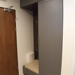 Szara szafa wnękowa z siedziskiem i złotymi uchwytami, zamontowana w przedpokoju obok brązowych drzwi.