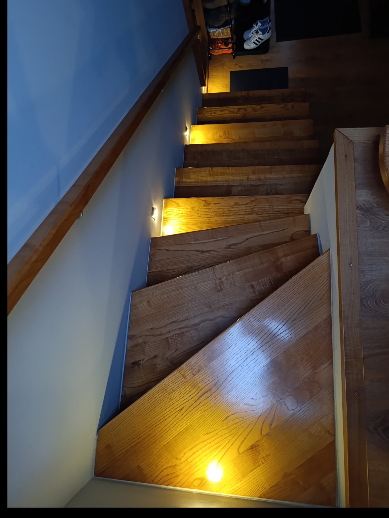 Drewniane schody z podświetleniem LED w ścianie, widok z góry, na górze schodów widoczna półka na buty i obuwie.