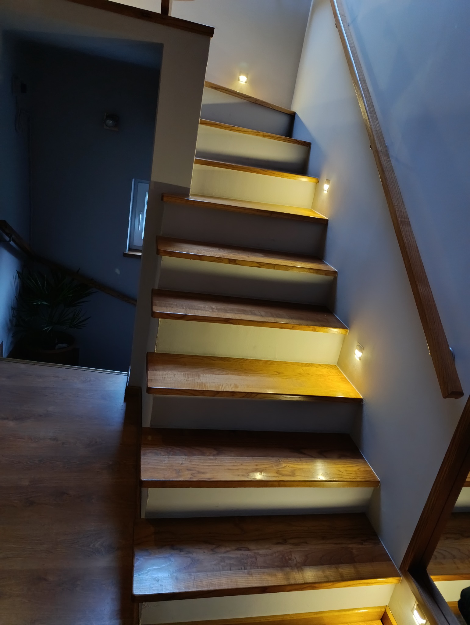 Nowoczesne schody drewniane z podświetleniem LED w ścianie, widok z dołu, jasne drewno i białe podstopnice, drewniana poręcz.