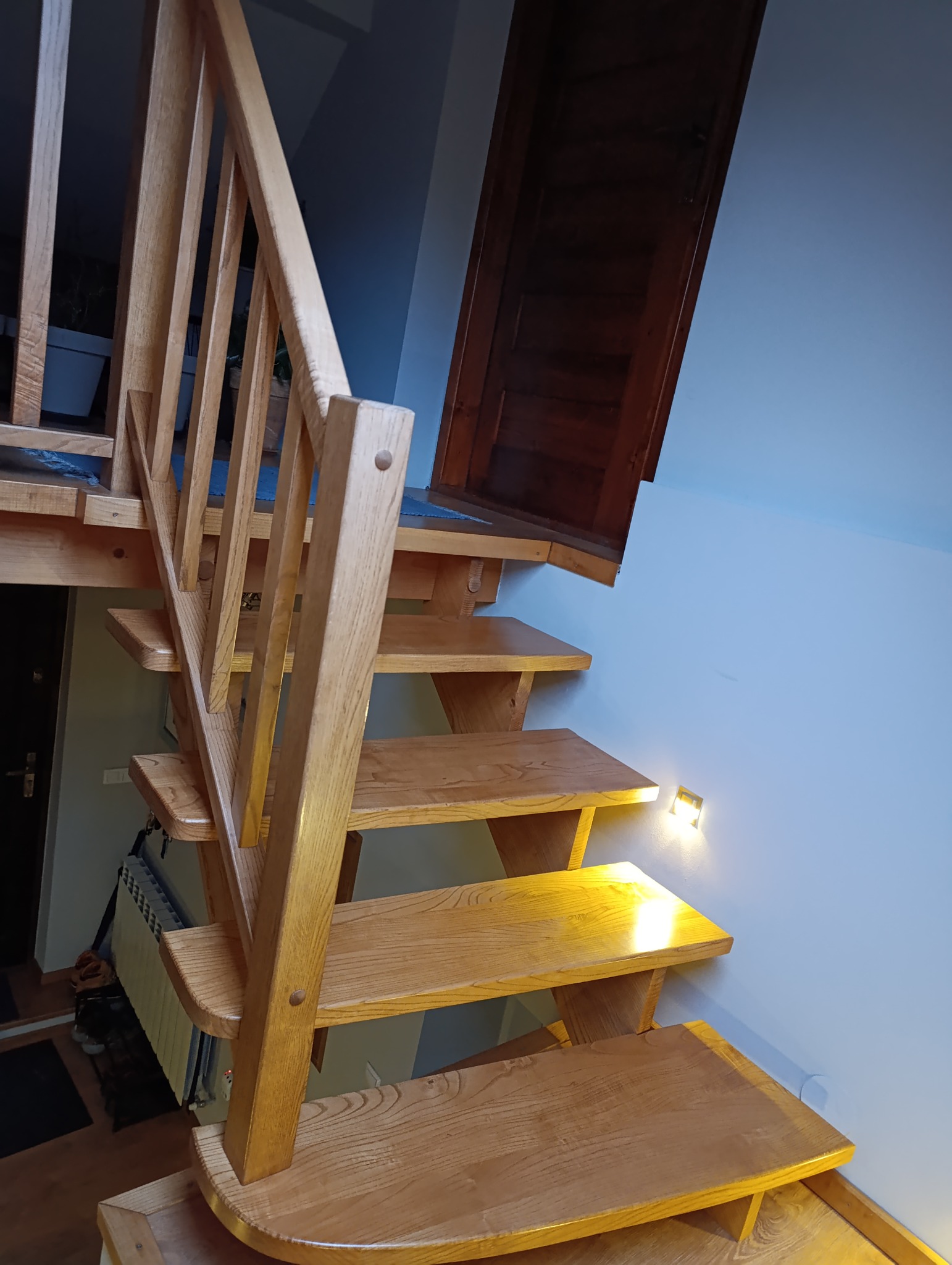 Drewniane schody z balustradą, oświetlone małymi lampkami ściennymi, prowadzące na antresolę z drewnianymi drzwiami.