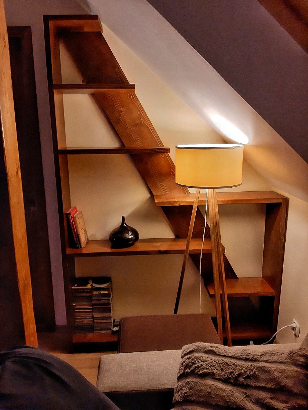 Drewniana konstrukcja regałowa w kształcie schodów, umieszczona w pokoju na poddaszu, oświetlona lampą stojącą z abażurem, z książkami i dekoracjami na półkach.