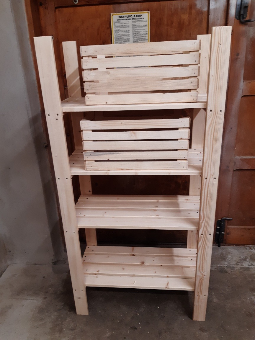 Surowa, drewniana półka z kilkoma poziomami, wykonana z jasnego drewna, z dwoma drewnianymi skrzynkami na górnych półkach, na tle brązowych drzwi i betonowej podłogi.