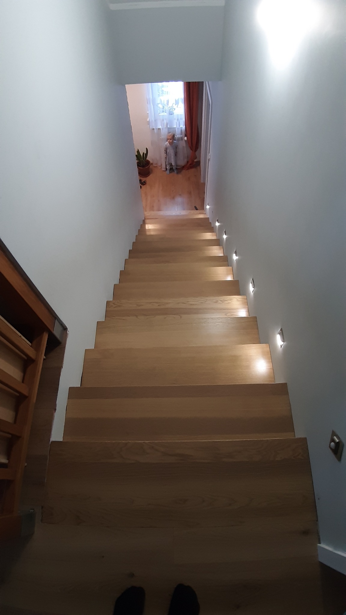 Widok z góry na drewniane schody z subtelnym oświetleniem punktowym wzdłuż ściany, w tle widoczna bawiąca się dziewczynka w pokoju.