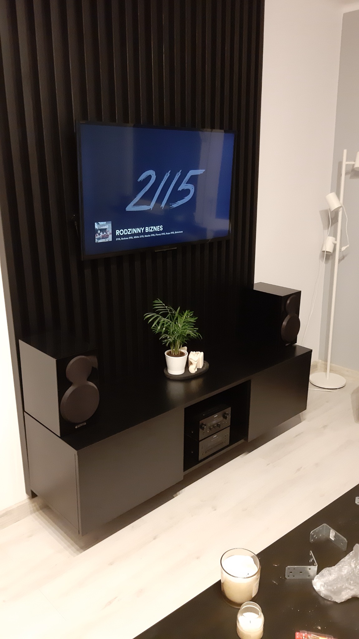 Czarna szafka RTV z drewnianymi lamelami na ścianie za telewizorem, głośniki po bokach, roślina doniczkowa na blacie, widoczne elementy montażowe na stoliku kawowym w tle.