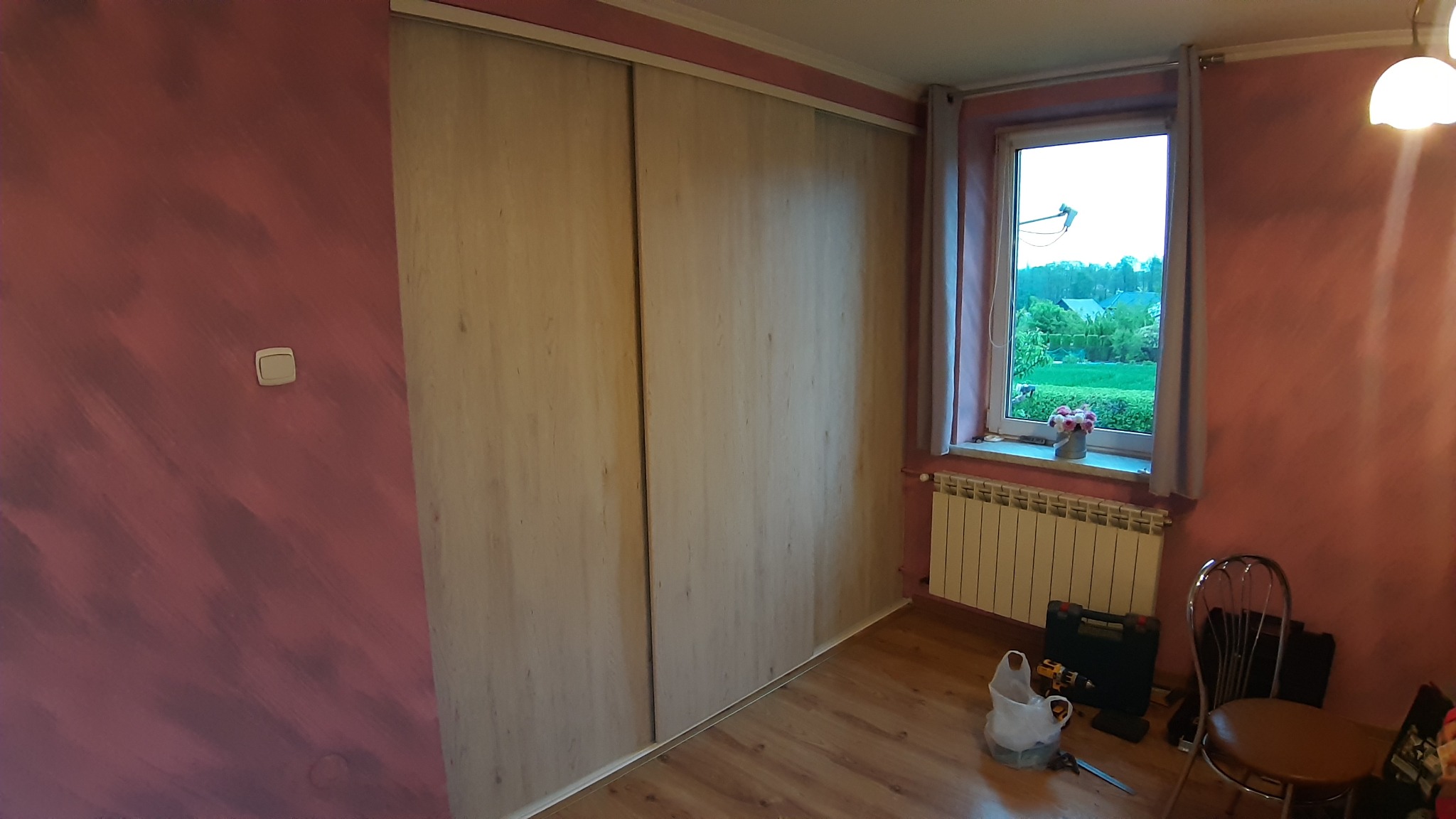 Jasna szafa wnękowa z przesuwnymi drzwiami, widoczna w pokoju z różowymi ścianami, narzędzia do montażu leżące na podłodze obok okna.