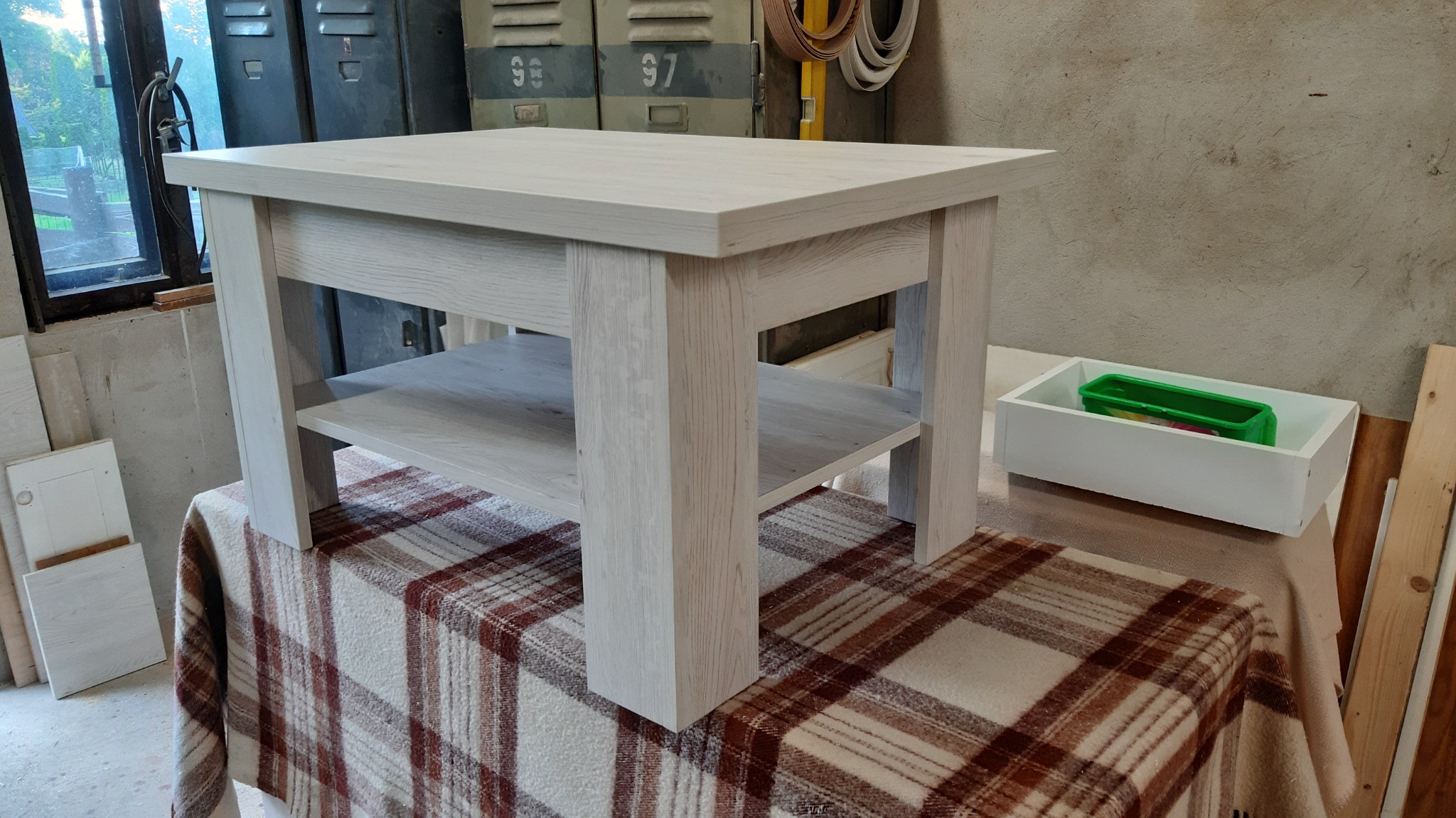 Stolik kawowy z jasnego drewna o widocznej strukturze, z dodatkową półką, ustawiony na pledzie w kratę w warsztacie.
