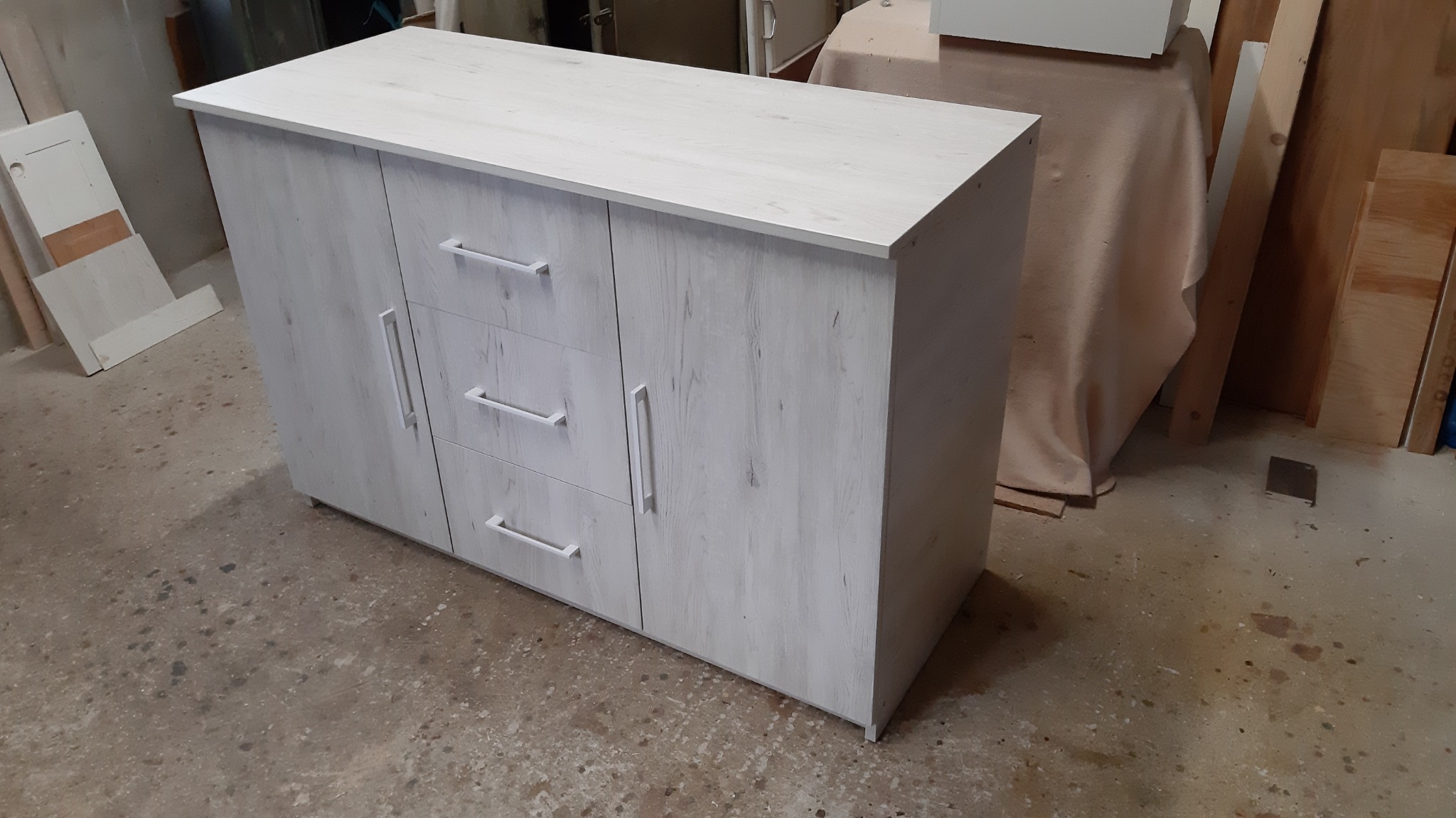 Biała komoda z trzema szufladami i dwojgiem drzwi, z podłużnymi uchwytami, stojąca w warsztacie na betonowej podłodze.