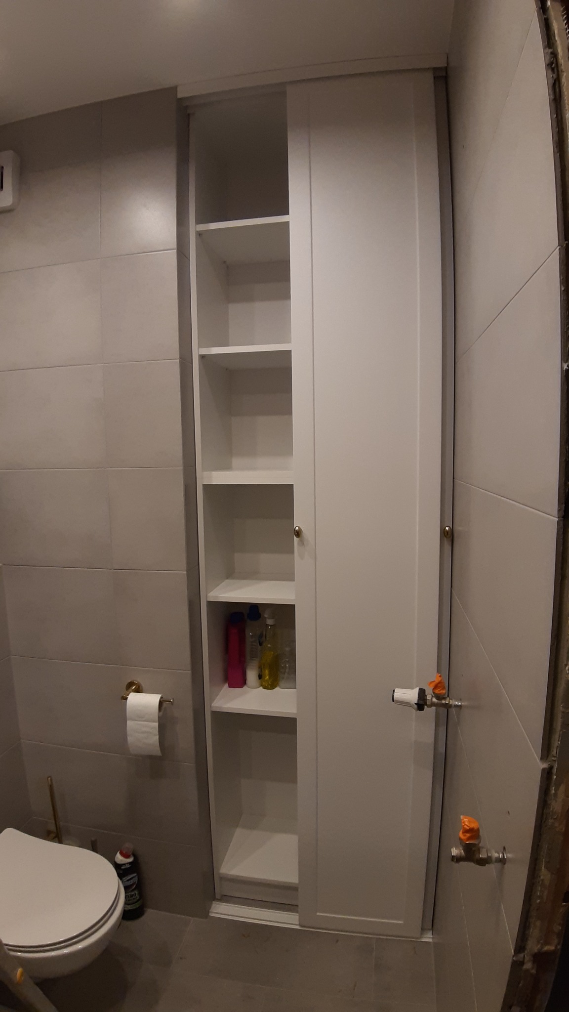 Biała szafa wnękowa z przesuwnymi drzwiami w łazience, widoczne półki z kosmetykami i środkami czystości, szare płytki na ścianach, toaleta w rogu.