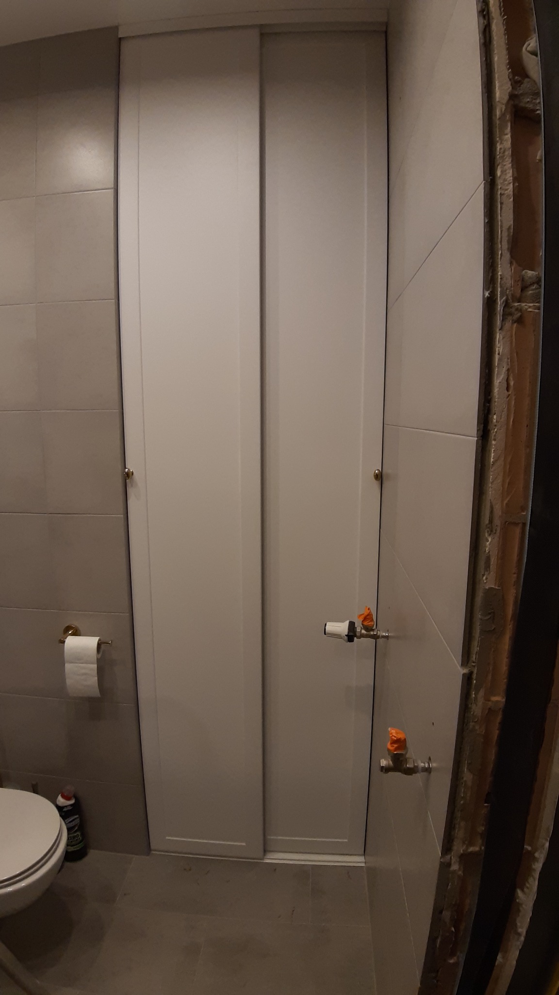 Białe drzwi przesuwne w łazience z szarymi płytkami, widoczne rury i zawory, oraz fragment ściany w trakcie remontu.