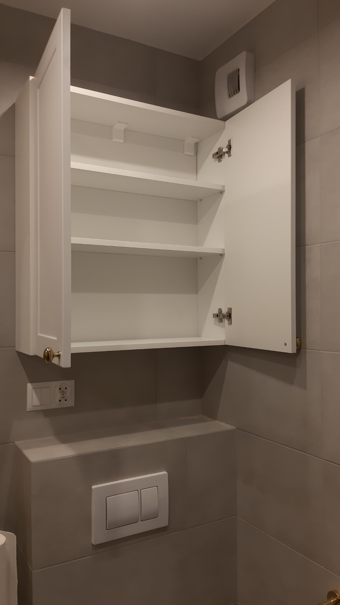 Biała szafka łazienkowa wisząca z otwartymi drzwiczkami, widoczne trzy półki, złoty uchwyt, szare płytki na ścianie i panelu z przyciskiem spłuczki