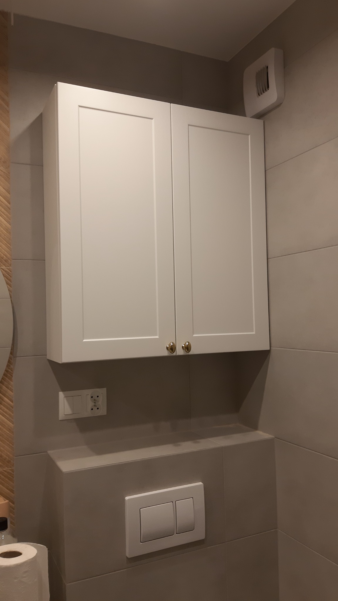 Biała szafka wisząca z dwoma drzwiczkami i złotymi gałkami w łazience z szarymi płytkami, nad zabudowaną spłuczką WC i gniazdkiem elektrycznym.